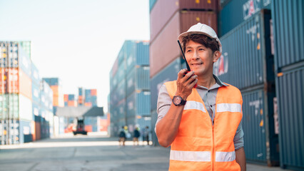 Asian worker safety vest hard hat walkie talkie shipping container cargo port logistics male outdoor professional worker smiling confidently during communication at busy cargo port yard © chokniti