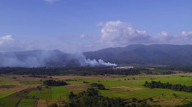 Air pollition releasing from factory buildings at the feet of large mountain formation, with lush tropical foliage and sunny clear sky in the background.