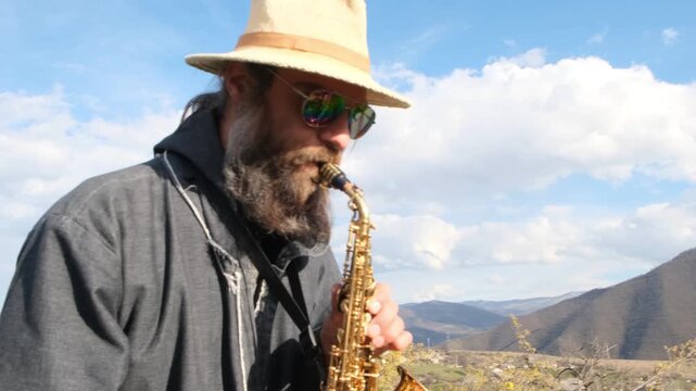 Tall slim trumpeter musician in hat, with beard and braid wearing sunglasses playing golden saxophone in mountain landscape against blue sky. Live music performance in nature. mysterious musician.