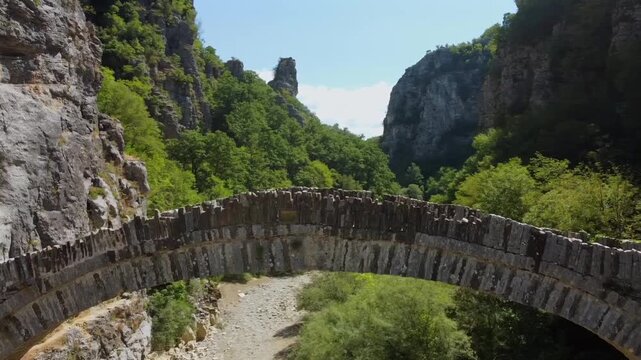 Cinematic drone pull-back shot revealing the historic Kokkoris arched stone bridge in Zagori. High-angle view of the traditional architecture and rugged Vikos Gorge landscape. No audio included.
