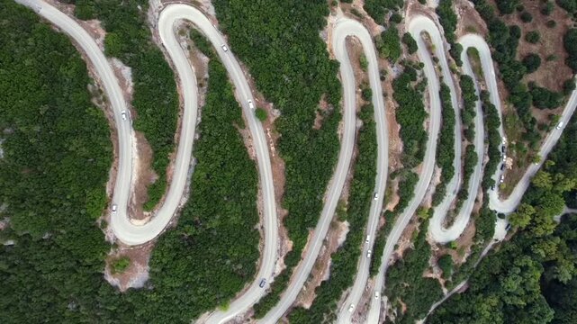 Cinematic top-down drone ascending shot of the famous zig-zag road in Papigo, Greece. High-angle view of white cars navigating the sharp hairpin turns through a lush forest. Ideal for travel films.