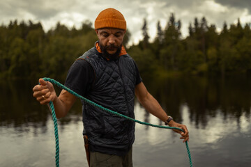 Man with a beard and an orange knit cap, wearing a quilted vest over a t-shirt, stands outdoors holding and looking down at a blue-green rope, with a lake and dark forest in the background. © Francis