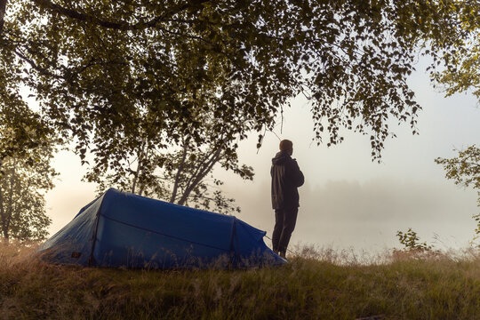 Lone person in silhouette stands by a blue tunnel tent overlooking a misty, golden-lit lake at dawn, framed by tree branches. Peaceful Nordic nature camping scene with warm, hazy light.