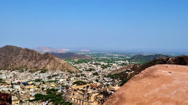 A wide aerial-style  showing the city spread between Aravalli hills with Nahargarh Fort walls on the ridges, dense buildings in the valley, and a clear blue sky viewed from the fort ramparts.