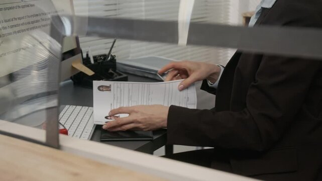 Medium close-up of anonymous female embassy officer in black formal suit jacket checking visa application and photos, verifying personal information in form, while working at desk with computer
