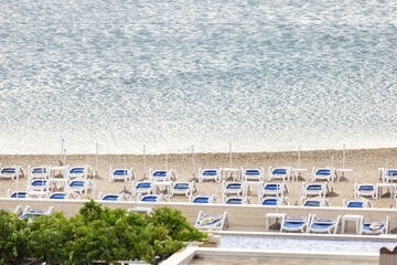 Fototapeta premium Rows loungers stand unused quiet seaside resort beach beside calm water pale sand, symbolizing tourism crisis weak season. Empty coastline still sea reflect decline of travel demand
