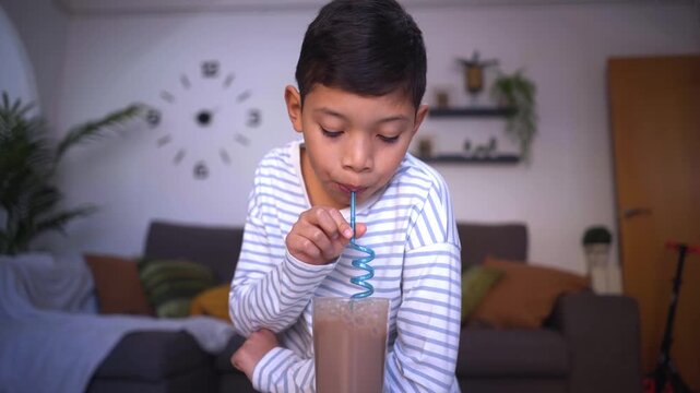 Young boy drinking chocolate milk with a straw at home in a cozy living room. Concept of childhood, everyday life and relaxed family routine