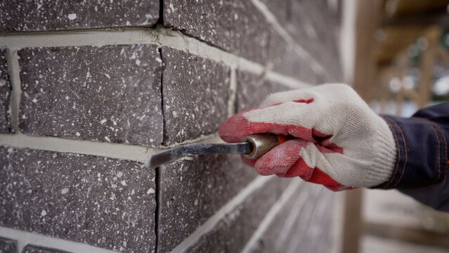 Worker's hand in a glove finishing mortar joints on a dark brick wall.
