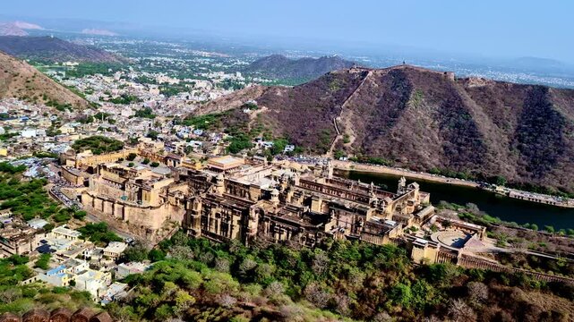 A high-angle  showing Amber Fort and Maota Lake below, with Nahargarh Fort walls stretching across the Aravalli hills and the city spread in the background under clear daylight.