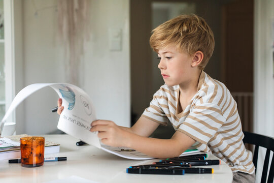 Boy with blond hair turning pages of drawing book while sitting at table