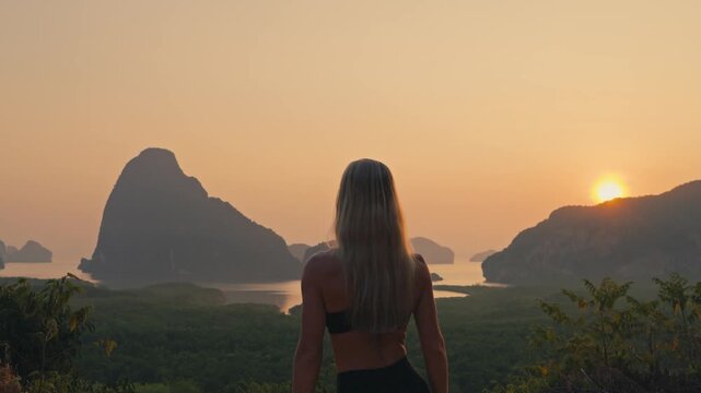 Fit blonde woman walks into frame and shields her eyes from the sun while gazing over a beautiful bay with towering islands. Warm golden sky creates a peaceful travel lifestyle scene