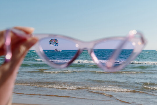 A creative POV shot of a hand holding pink sunglasses to frame a sharp view of a parasailer over the blue ocean. This image captures the essence of summer travel, vacation leisure at a coastal resort.