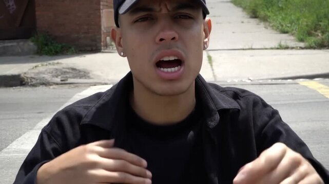 Tracking shot of young man rapping and gesturing on urban sidewalk curb in daytime neighborhood with buildings and pavement