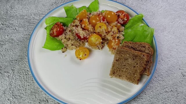 Whole grain bread and salad with quinoa, cherry tomatoes, and lettuce is served on a white plate on a gray table. Healthy eating concept. Lenten dish