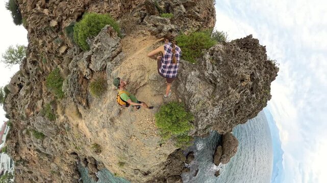 A Man and Woman stand on a rocky cliff overlooking the sea, surrounded by rugged terrain. The scene is captured with a tiny planet effect, creating a spherical view of the landscape.