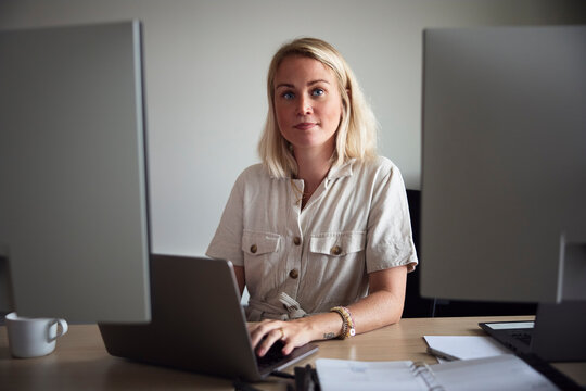 Portrait of blond young female tech professional sitting with laptop in office desk