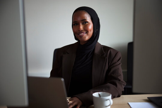 Portrait of smiling female business entrepreneur wearing headscarf sitting at desk in office
