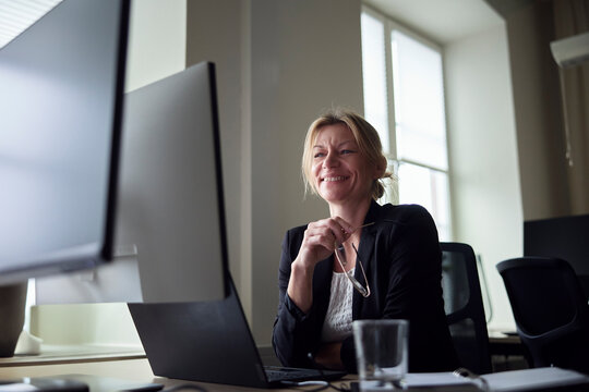 Low angle view of smiling female entrepreneur sitting with laptop and computer at office desk