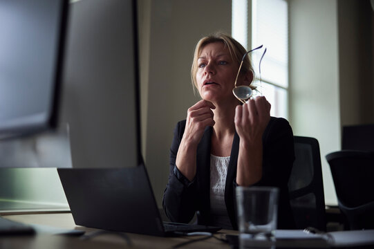 Mature female tech expert holding eyeglasses looking at computer while sitting at office desk