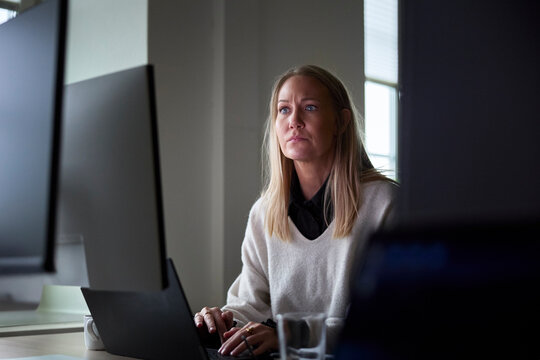 Concentrated female tech professional working on laptop and computer at desk in office