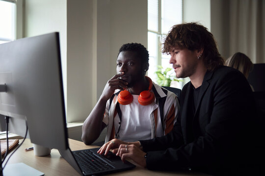 Male tech professional with wireless headphones around his neck sitting beside colleague using laptop in office