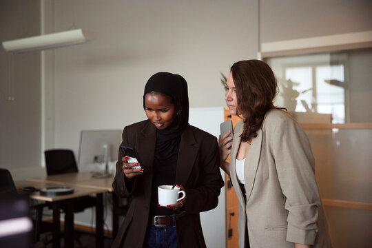 Businesswoman standing near female colleague using smart phone in office