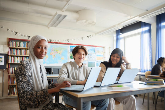 Portrait of smiling students sitting with laptops on desk in classroom