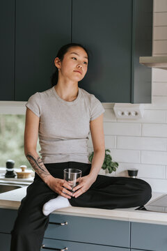 Contemplative young woman sitting with glass of water on kitchen counter in home