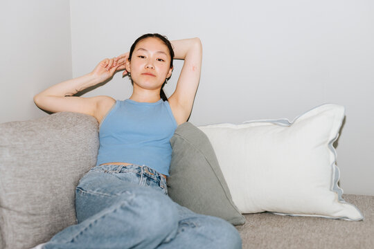 Portrait of beautiful young woman posing with hands behind head while sitting on sofa at home