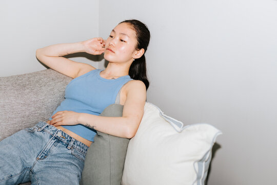 Thoughtful young woman leaning on wall while sitting at sofa in home