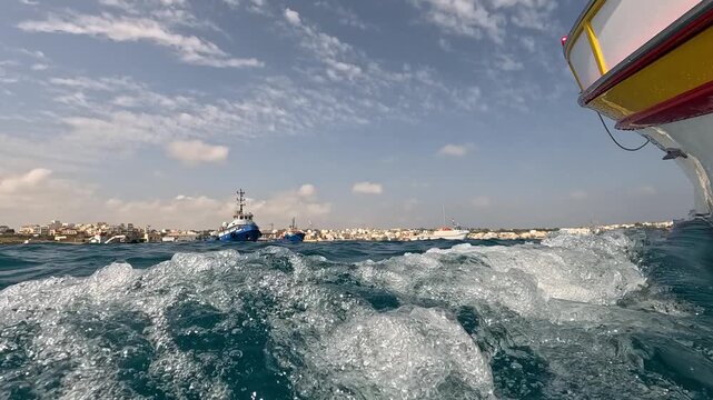 Low-angle shot from a moving boat capturing sea splashes with traditional fishing boats and coastal houses of Marsaxlokk village in the background
