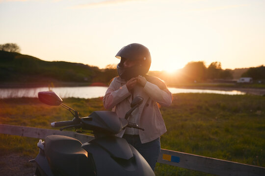 Girl fastening helmet while standing next to scooter against sky at sunset
