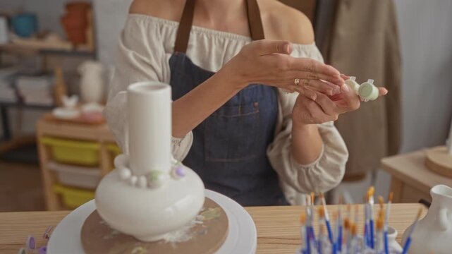Woman holding a row of small paint pots while gesturing with her hand over a ceramic vase in studio; serenity craft focus.