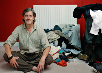 Man sitting crosslegged on bedroom floor with pile of clothes © colin
