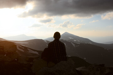 Fototapeta premium A tourist sits on the edge of a rock and enjoys the sunset on spring mountains with snowy peak on the background. Landscape photography