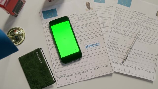 Top view flatlay close-up of European Union visa applications with blue approval stamp, smartphone with plain green chromakey screen, passport, on officer desk at foreign embassy