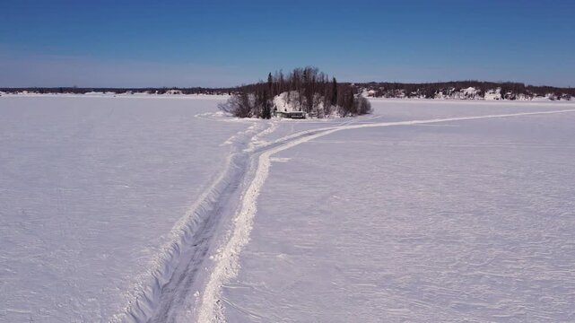 Aerial 4K 60fps low-level drone footage flying down plowed ice road toward island cabin, Big Lake, Alaska. Snow berms flank road leading directly to remote island home in trees.