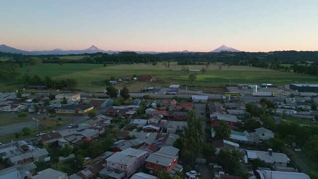 Forward flight over small town rooftops and green farmland with a row of snow-capped Andean volcanoes glowing at sunset. Entre Lagos, Chile