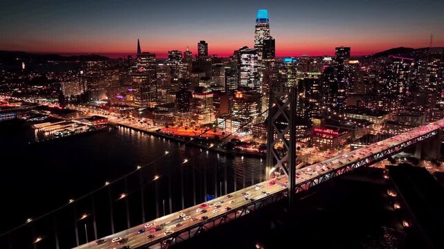 Aerial drone view of downtown San Francisco at dusk, featuring the Bay Bridge, waterfront, Salesforce Tower, glowing city lights, and a vivid sunset sky over San Francisco Bay.