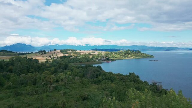 Low altitude flight over dense treetops revealing wide blue lake with golden farmland peninsula and distant islands under partly cloudy sky. Futrono, Chile