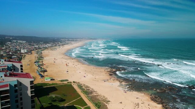 Reveal shot rising above coastal apartments to expose long golden beach with rolling Pacific waves and resort town stretching along the shoreline. El Tabo, Chile