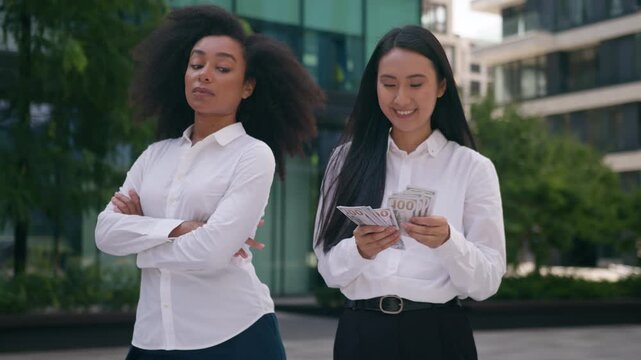 Unhappy Asian African American business women counting money outdoor jealous colleague crossed arms financial rivalry salary bonus income discrimination businesswomen girls cash pay rich envy conflict