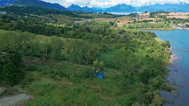 Reveal shot over dense forest with isolated blue cabin on lake shore unveiling vast turquoise lake and Andean mountain range beyond. Futrono, Chile