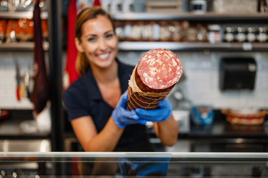 A happy female deli worker wearing blue gloves presents a large cured meat sausage at the counter of a butcher shop.