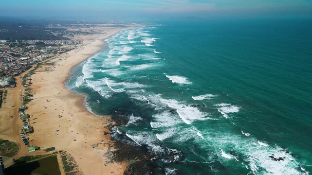 Reveal shot over sweeping endless golden beach with parallel rows of turquoise Pacific surf and coastal town stretching to the misty horizon. El Tabo, Chile