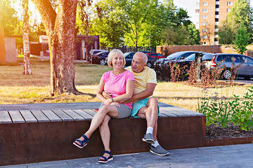 Smiling older man and woman sit together on wooden bench in sunny urban park, casual colorful clothes, greenery and trees, warm summer mood
