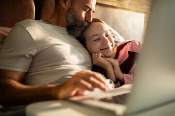 Father kissing daughter while cuddling in bed with laptop at home