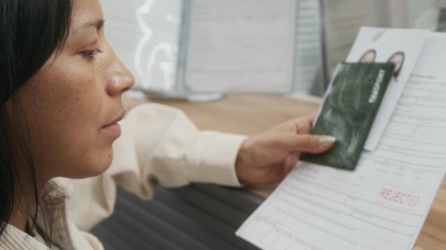 Medium close-up of disappointed middle-aged Hispanic woman receiving rejected tourist or immigration visa application from anonymous consular officer, face expressing emotions of dismay and failure