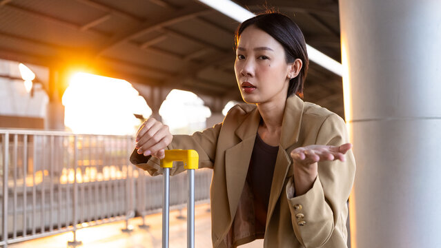 Confused Asian woman waiting for train at platform in morning sunrise. Holding yellow luggage, shrugging. Concept of delayed commute, missed trip, or travel problems.