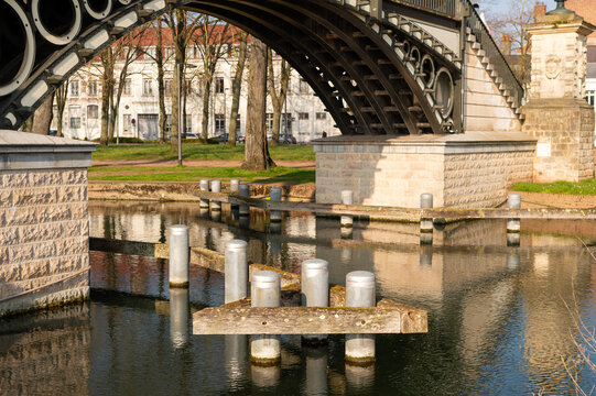 View under a cast iron arch bridge over a canal in Lille France featuring stone pillars and wooden water barriers.